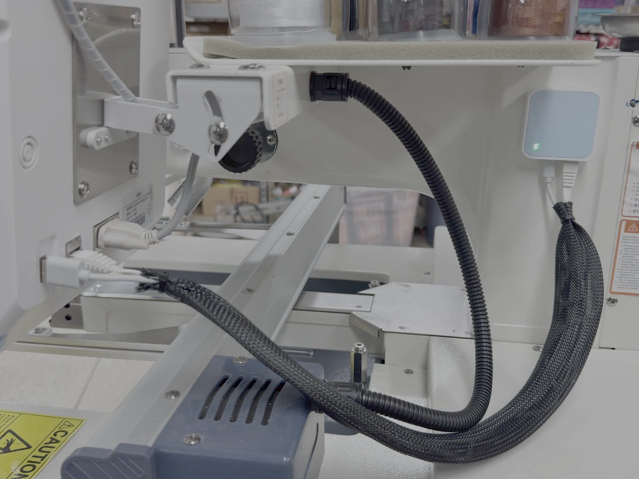 The Wi-Fi bridge mounted on the side of an embroidery machine, its power and network cables running in a sleeve to the machine's USB port and network socket